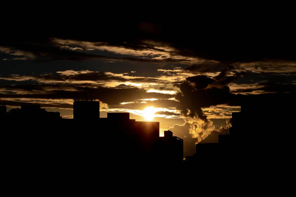 Sol baixo entre silhuetas de prédios sob um céu de nuvens escuras